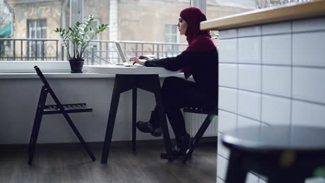 piękna muzułmańska dziewczyna z hidżabem na głowie, siedząca w pobliżu okna, pisze coś na klawiaturze laptopa