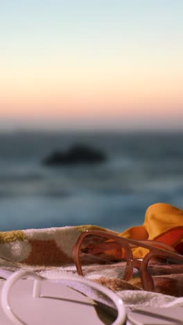 Video-Vertical-Concepto-De-Vacaciones-De-Verano-De-Toalla-De-Playa-Con-Chanclas,-Gafas-De-Sol-Y-Sombrero-Para-El-Sol-Contra-El-Cielo-Del-Atardecer