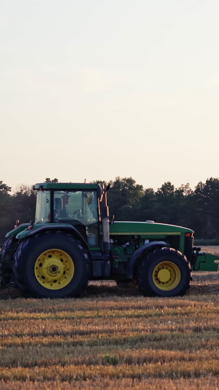 Agricultural machine pressing hay on the golden field. Harvesting dry grass for livestock. Green and red tractor during seasonal works in summer. Vertical video