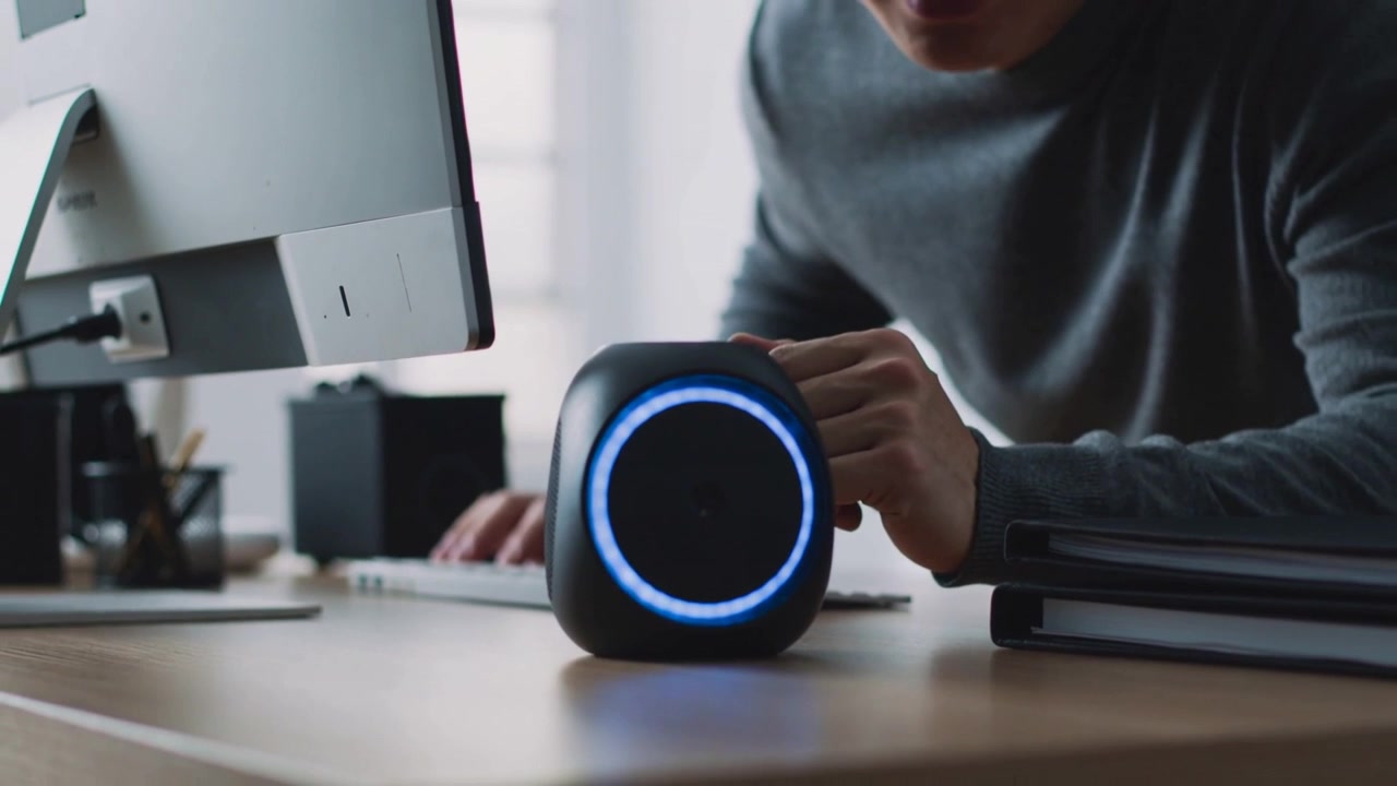 Anello LED blu luminoso, un impiegato con maglia grigia si sporge, allungando la mano verso lo smart speaker per parlare.