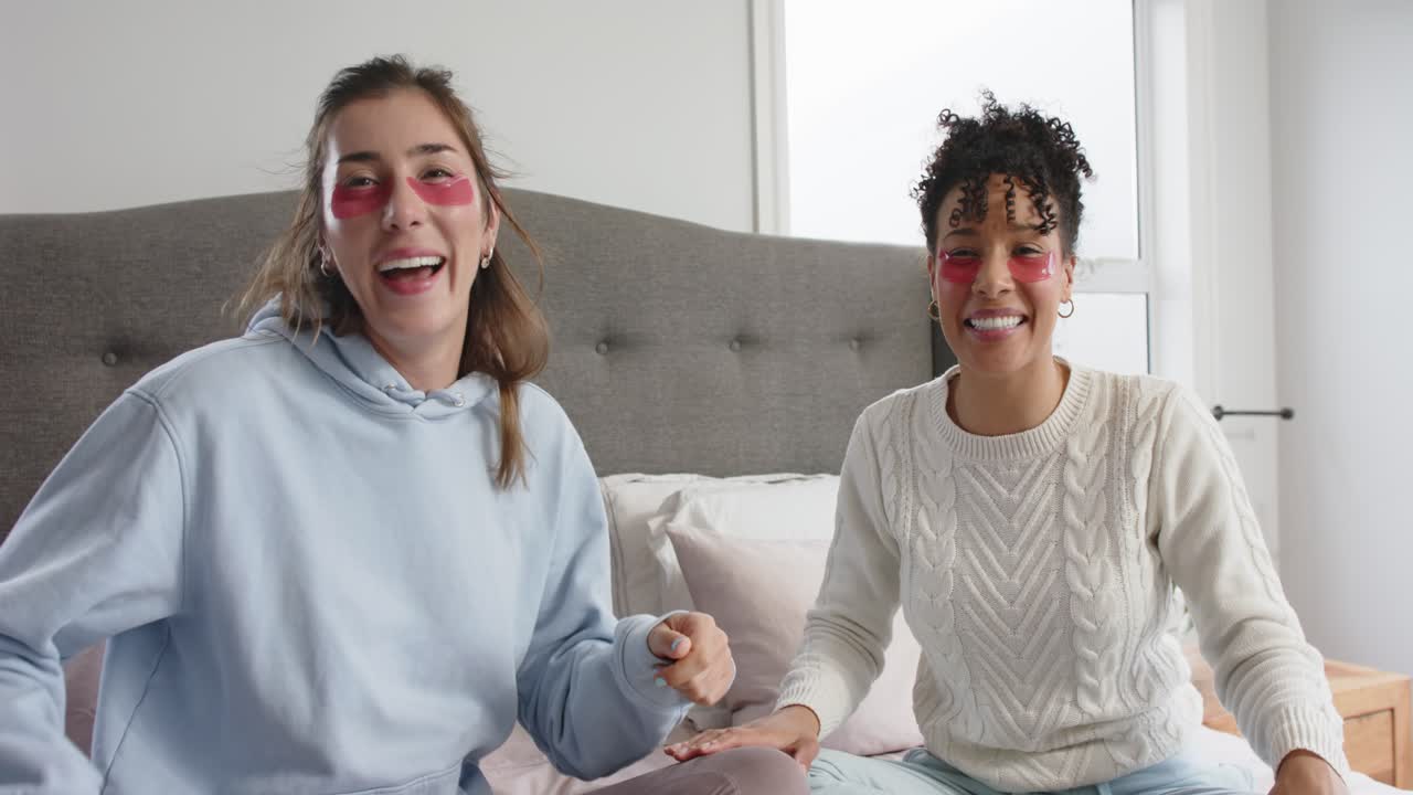 Amiche diverse che ridacchiano sollevando e applicando patch occhi in gel rossi per rinfrescare gli occhi sul letto.