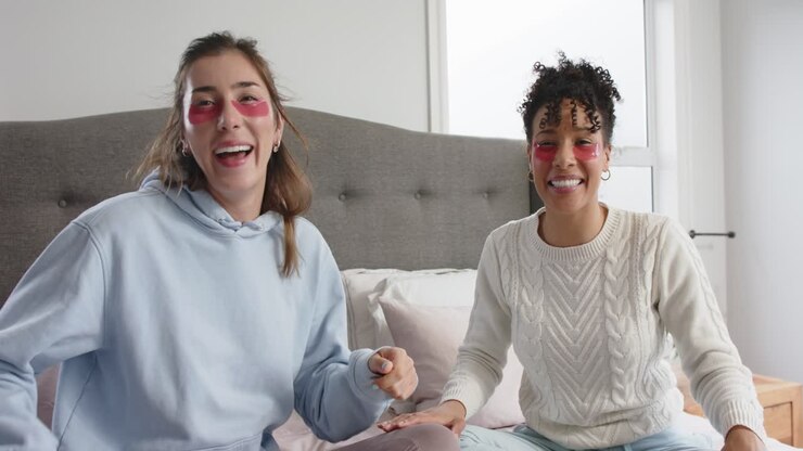 Amiche diverse che ridacchiano sollevando e applicando patch occhi in gel rossi per rinfrescare gli occhi sul letto.