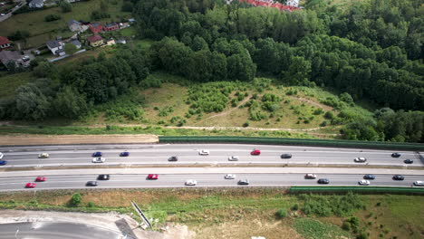 Luftaufnahme-Aus-Großer-Höhe-Des-Fließenden-Autosverkehrs-Auf-Einer-Mehrspurigen-Landstraße,-Der-Sich-Auf-Beiden-Seiten-Der-Straße-Bewegt,-Umgeben-Von-Feldern,-Grünen-Feldern,-Bäumen-Und-Vorstadthäusern,-Reisen-Und-Transport