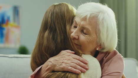 close up of the gray haired caucasian woman smiling and hugging her cute blond teenager granddaughter while they sitting on the couch in the living room. portrait. inside.