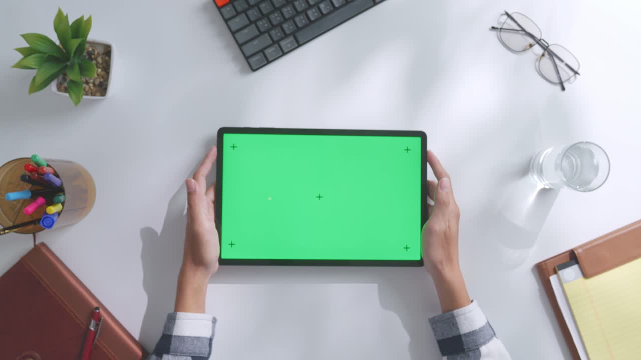 верхний вид ноутбука с макетным зеленым экраном chromakey на деревянном офисном столе рядом с ноутбуком с ручками, очками и стаканом воды.