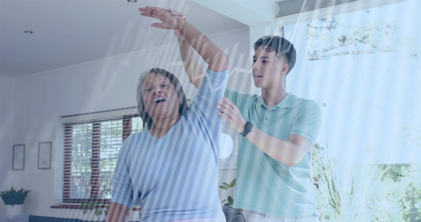 Người chăm sóc đặt tay dưới cánh tay người phụ nữ lớn tuổi, hướng dẫn nâng người bằng thiết bị treo trên cao để phục hồi chức năng, rèm cửa có thể nhìn thấy.