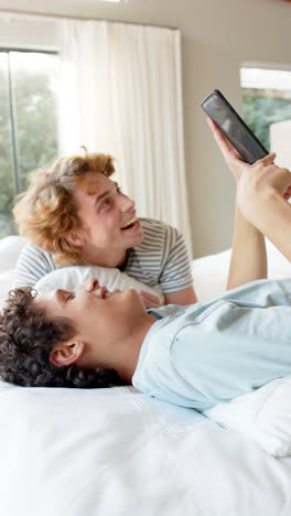 vertical video of happy diverse gay male couple lying on bed using smartphone at home, slow motion