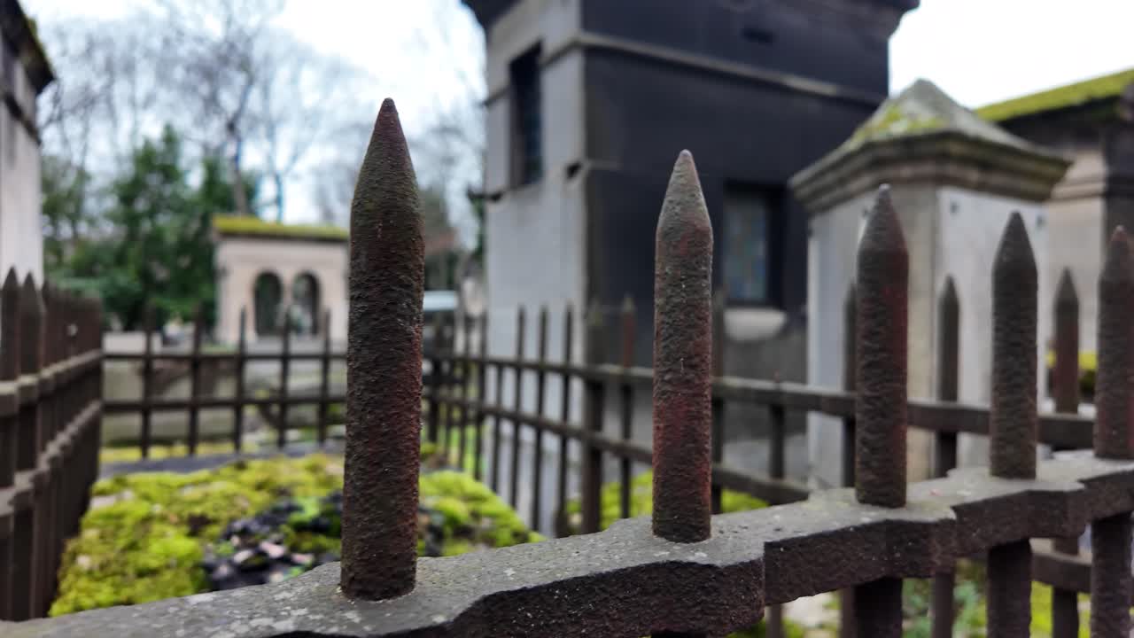 Primo piano di una recinzione rustica con punte di ferro che protegge tombe ricoperte di muschio e mausolei logori in un vecchio cimitero