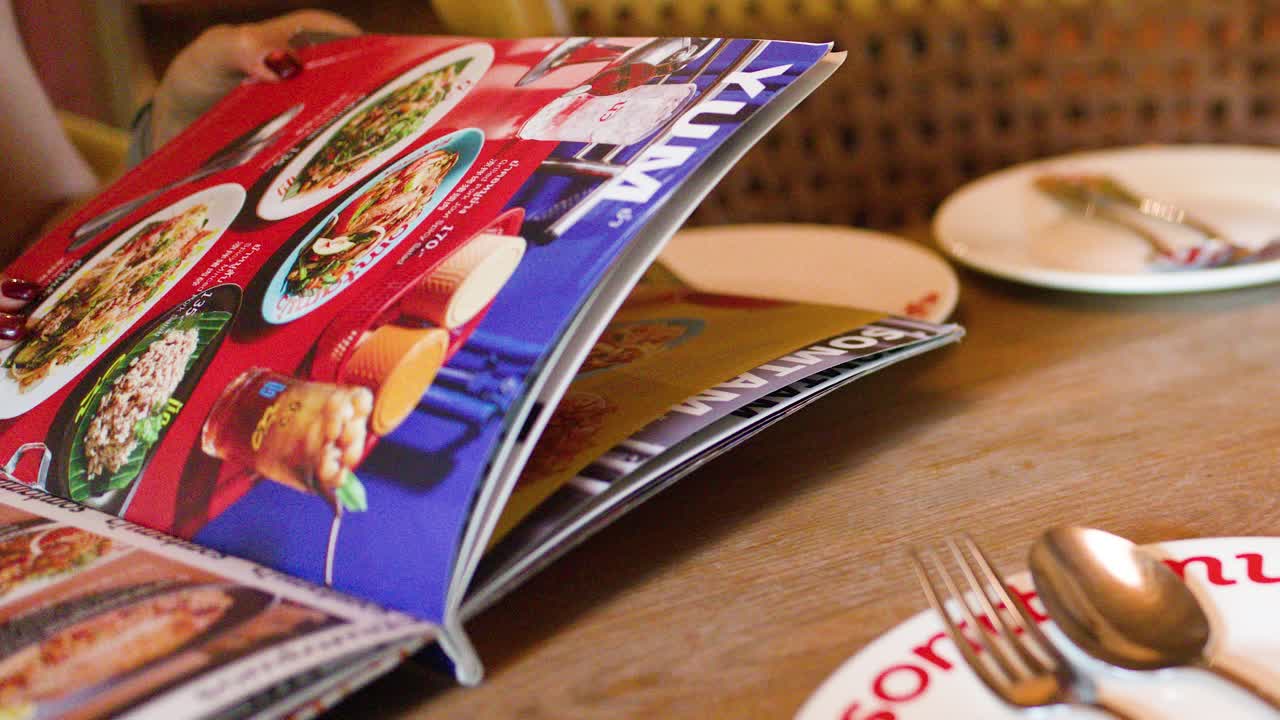 Hand flips through vibrant Thai food menu at sunlit restaurant table, utensils and plates visible