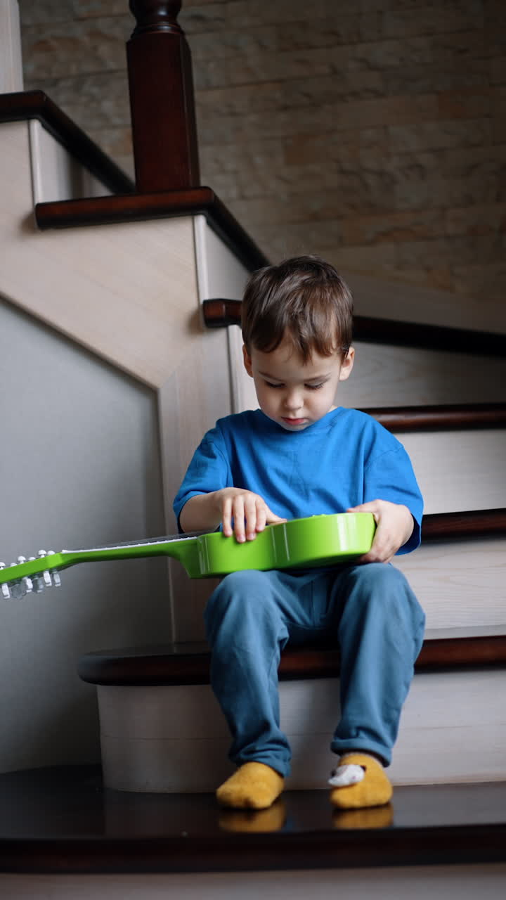 Small kid sits on the stairs being busy with his guitar. Portrait of a toddler focused on his toy. Vertical video.