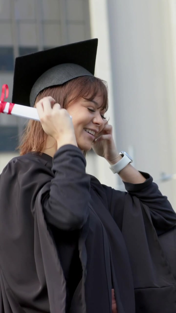Video verticale: Donna in toga nera che tiene una pergamena, alza il tocco e si sistema i capelli nel campus per una cerimonia