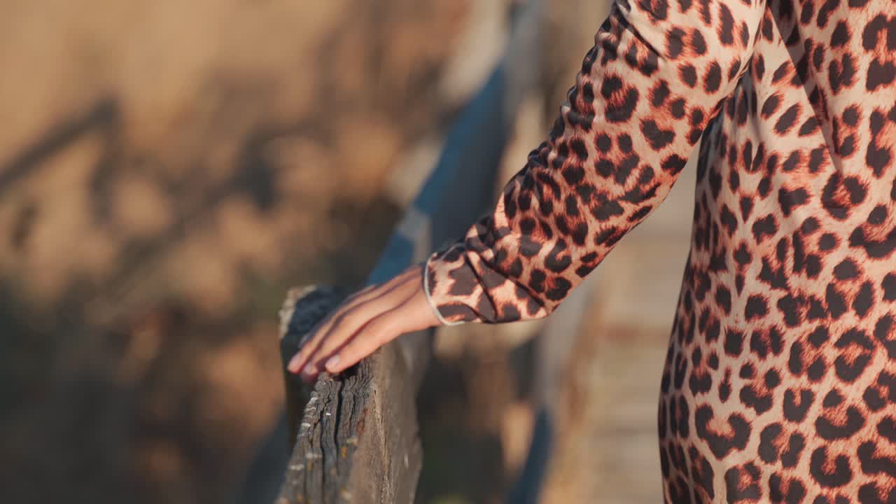 Caucasian woman wearing animal printed dress walking on pathway gliding hand across outdoor wooden handrail in countryside, handheld behind close up