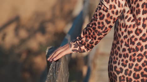 caucasian woman wearing animal printed dress walking on pathway gliding hand across outdoor wooden handrail in countryside, handheld behind close up