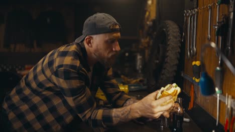 side view of a mechanic guy in a plaid shirt drinking soda and eating a burger during lunch in a studio workshop