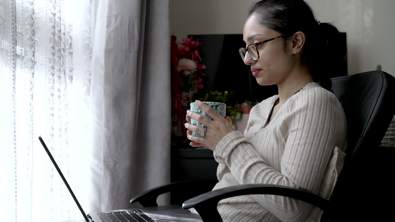 한 사람이 창 에 앉아서 을 마시면서 노트북에서 일하면서 다중 작업을 하는데, 이는 디지털 미디어 사용과 가정의 일상 생활의 원활한 통합을 강조합니다.