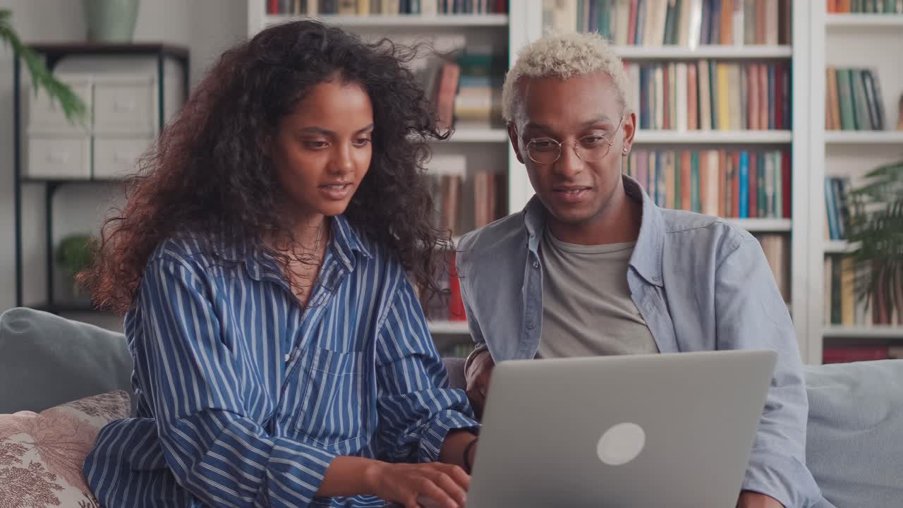 Mixed ethnic couple sit relax on couch in living room choose new goods on laptop
