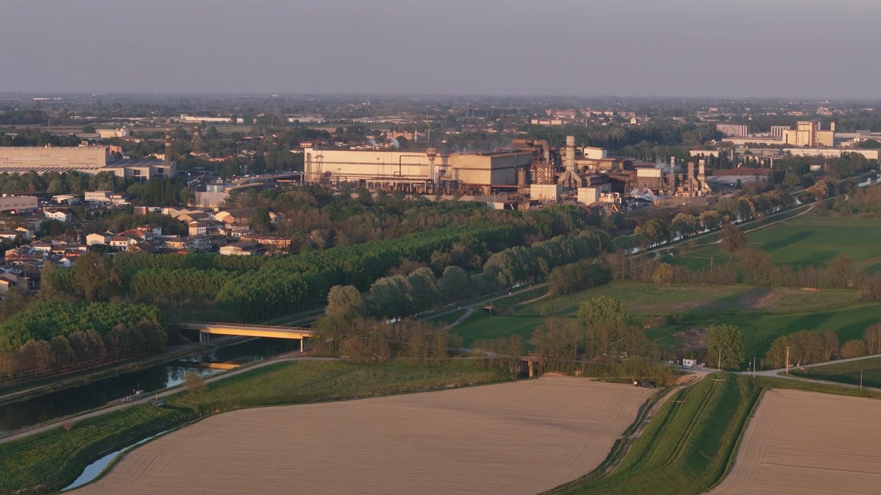 Complesso siderurgico industriale che si estende lungo il canale navigabile mentre la pianura agricola circostante della Valle del Po si espande all'orizzonte, movimento aereo lento