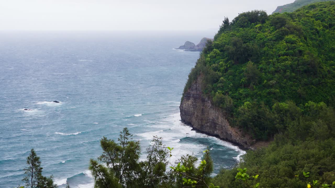 A high overlook frames a lush sea cliff above slate water as white surf bands sweep the shoreline, while scattered islets blur in haze and trade winds ruffle the surface