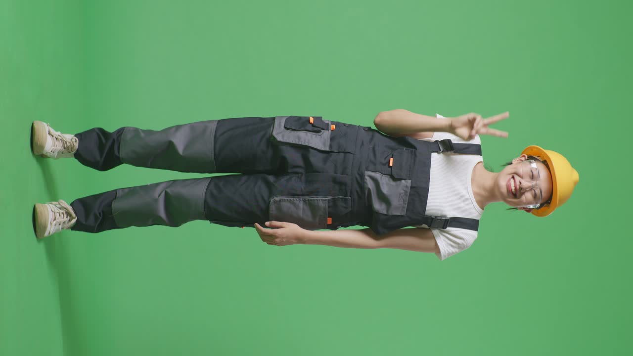 Full Body Of Asian Woman Worker Wearing Goggles And Safety Helmet Smiling And Showing Peace Gesture To Camera While Standing In The Green Screen Background Studio