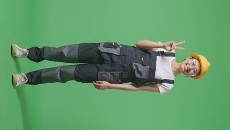 full body of asian woman worker wearing goggles and safety helmet smiling and showing peace gesture to camera while standing in the green screen background studio