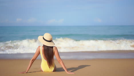 una mujer joven con un traje de baño amarillo de una pieza se sienta frente a las olas del océano mientras llegan a la orilla