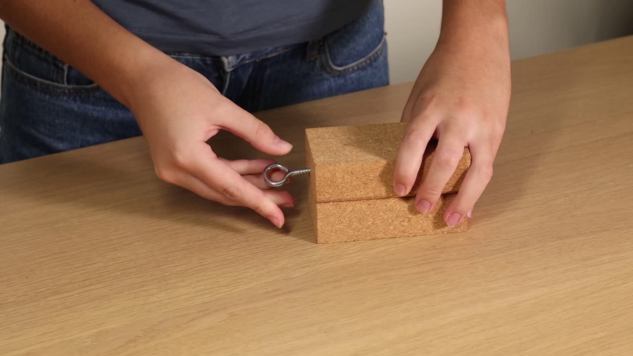 Hands manipulate a wooden block, inserting a hook screw on a table. Natural lighting highlights the detailed assembly process
