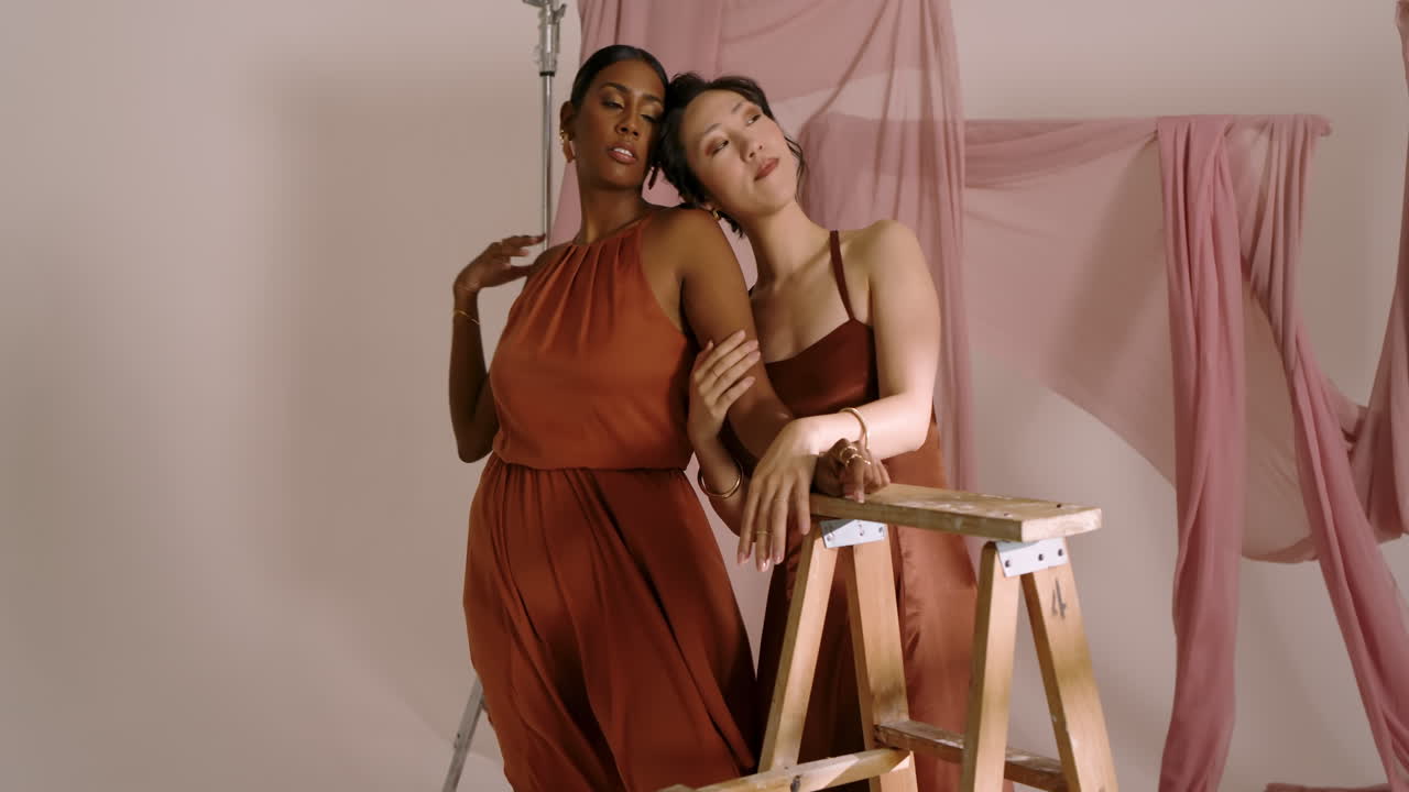 Diverse female colleagues exchanging glances then wrapping arms around ladder rung on pink backdrop