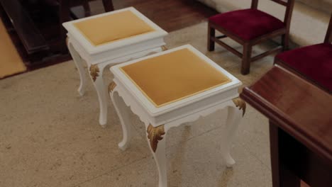 two ornate white stools with gold cushions, placed in a formal indoor setting with wooden chairs nearby