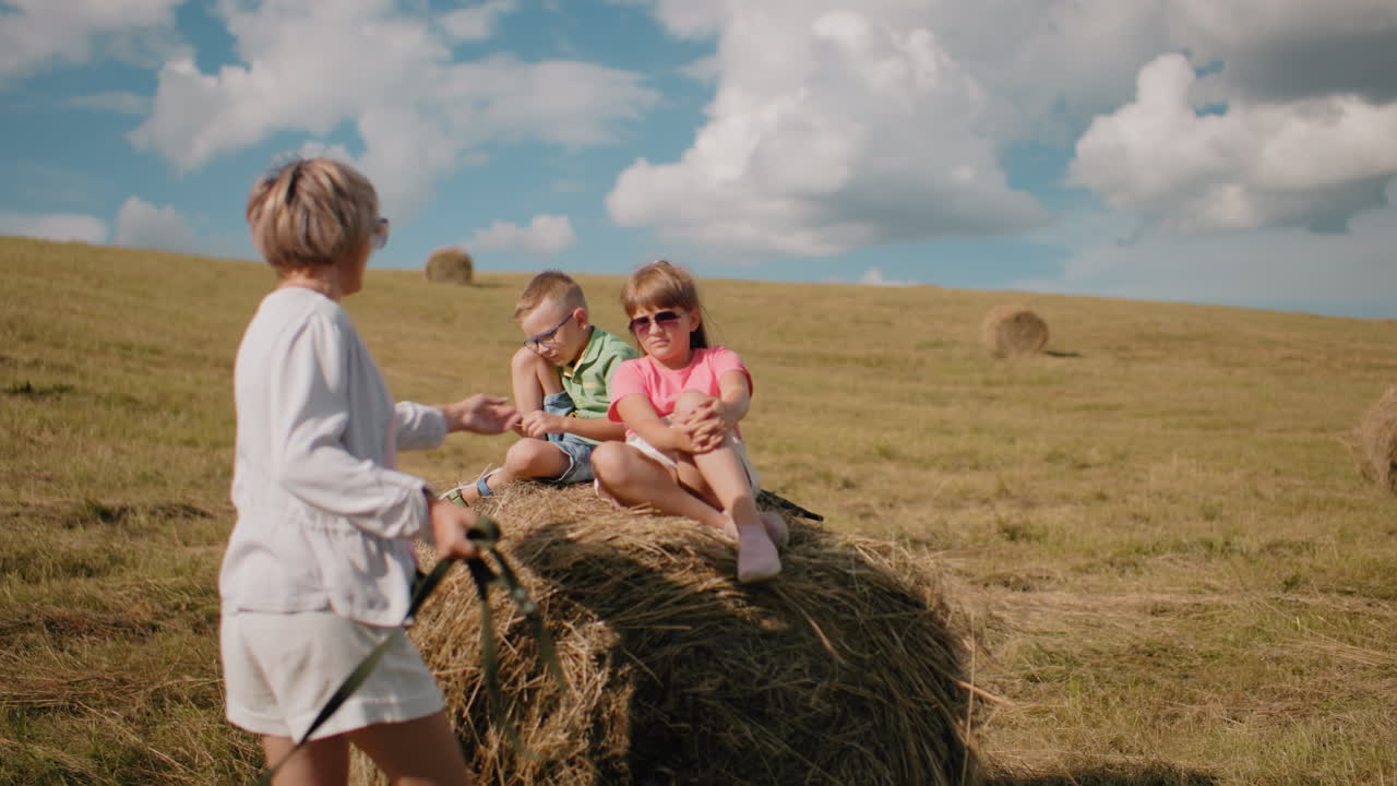 двое детей сидят на топке сена в солнечной сельской местности, девушка в розовом топке носит солнцезащитные очки, мальчик в зеленом корректирует шнурки, тетя подходит, чтобы помочь девушке вниз, открытое поле с рассеянными топками сена
