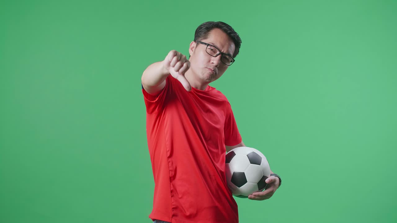 hombre asiático con una pelota mostrando los pulgares hacia abajo gesto para burlarse de los perdedores mientras anima el fútbol en el fondo de la pantalla verde