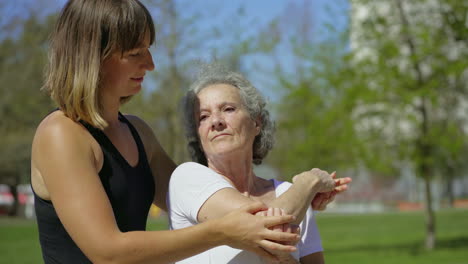 focused elderly lady with coach stretching arm in park .