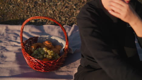 woman sits on plaid with basket of food. healthy snack in silence with nature by river at sunset. relaxation in solitude and reflection on existence