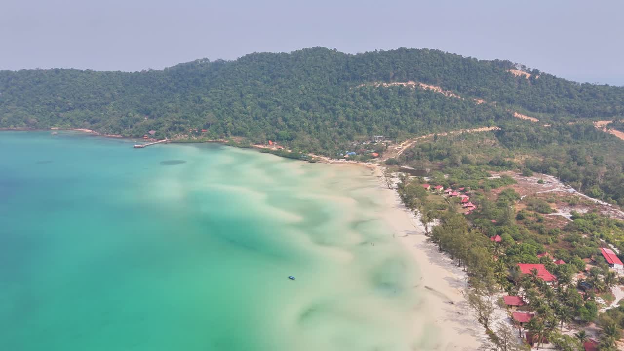 Aerial view of Kaoh Rong Sanloem in Cambodia with turquoise waters, a white sandy beach, lush green hills, and scattered red-roof houses along the shoreline, creating a serene tropical landscape