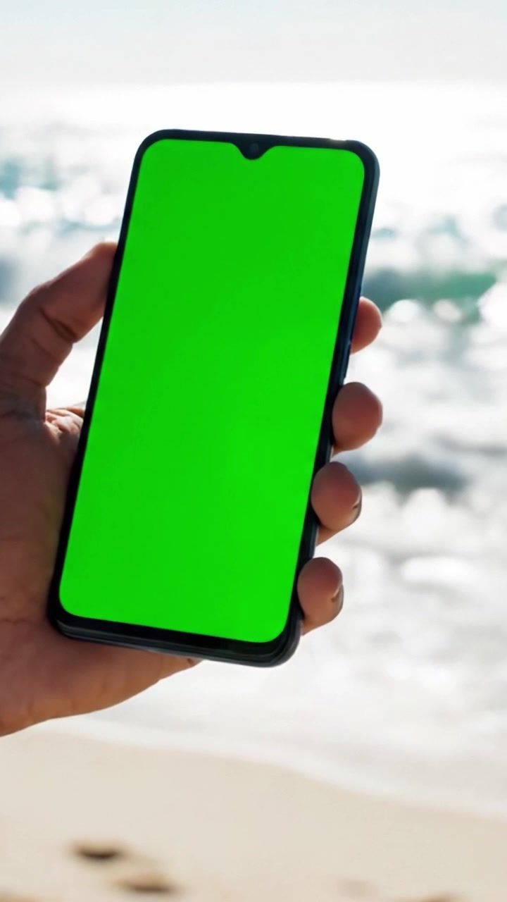 Una persona che tiene uno smartphone con uno schermo verde sulla spiaggia, mostrando il dispositivo contro uno splendido sfondo naturale.