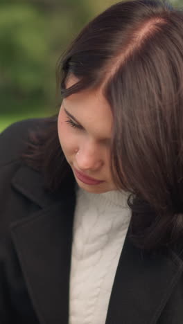 joven con abrigo negro y cuello gordo blanco centrada en el teléfono con el cabello revoloteando en el viento, sentada al aire libre en un parque con vegetación borrosa y un objeto poco claro en el fondo