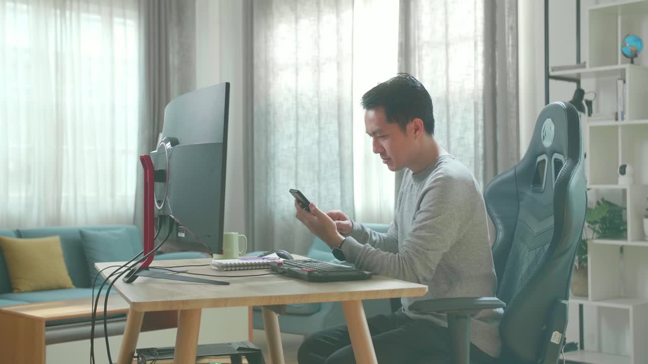 Asian Man In Long Sleeved T-Shirt And Black Pants Using Mobile Phone While Using Desktop Computer For Working At Home.