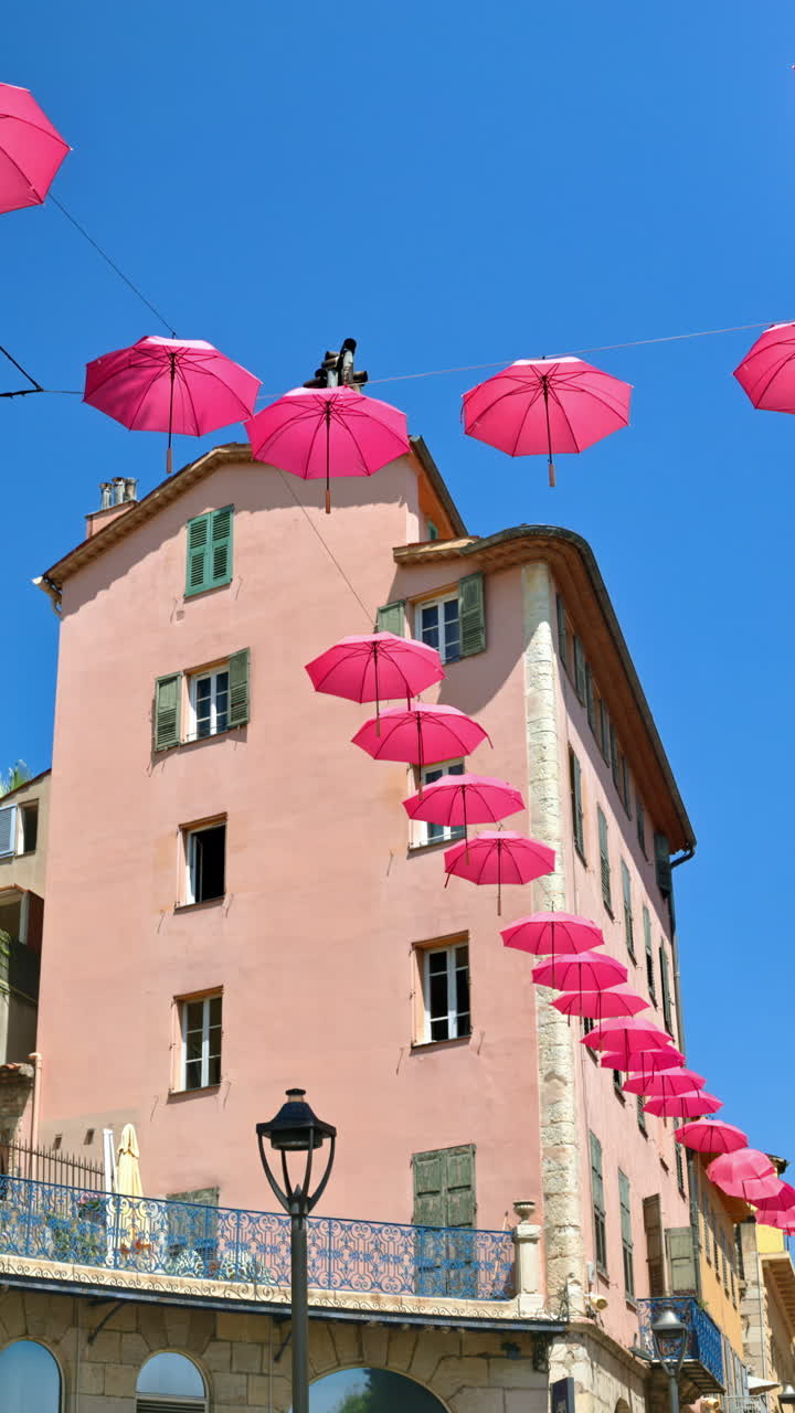 Rows of pink umbrellas above the streets of the old town in Grasse, France with a building on the background. Vertical