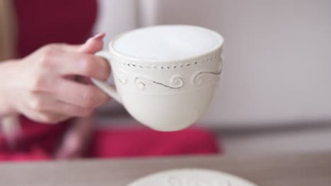 Moving-focus-from-nice-white-cup-with-cappuccino-to-young-woman-with-long-blonde-hair-sitting-at-the-table-and-relaxing-in-the