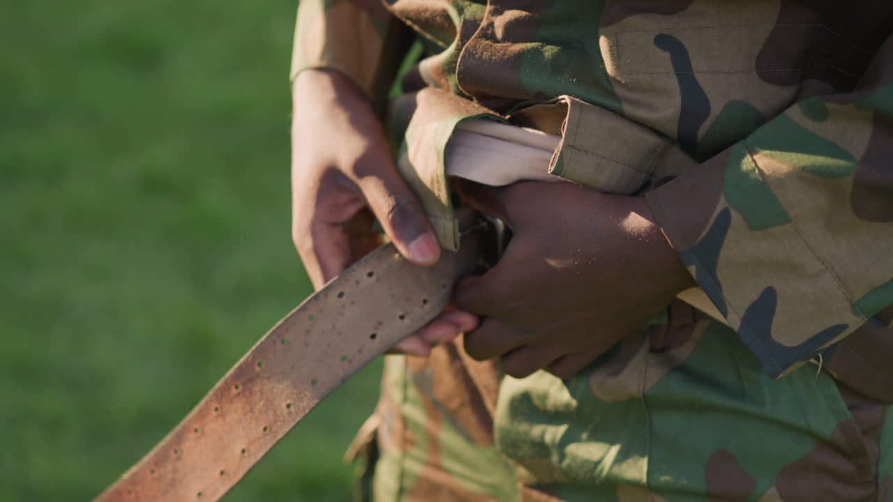 Nærbillede af soldat, der spænder læderbælte udendørs, mand der fastgør læderrem på militæruniform i sollys, detaljeret visning af soldat, der justerer sit bælte under udendørs træningssession
