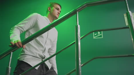 guy with serious face in white shirt and grey trousers walking up stairs with iron rail. green background adds contrast, while soft lighting creates a dramatic in modern indoor setting