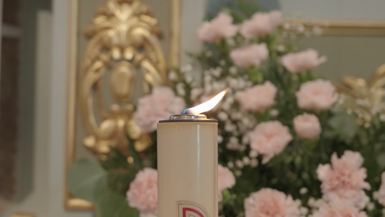 Vista cercana de una vela votiva encendida dentro de una iglesia con flores suaves y un altar dorado.