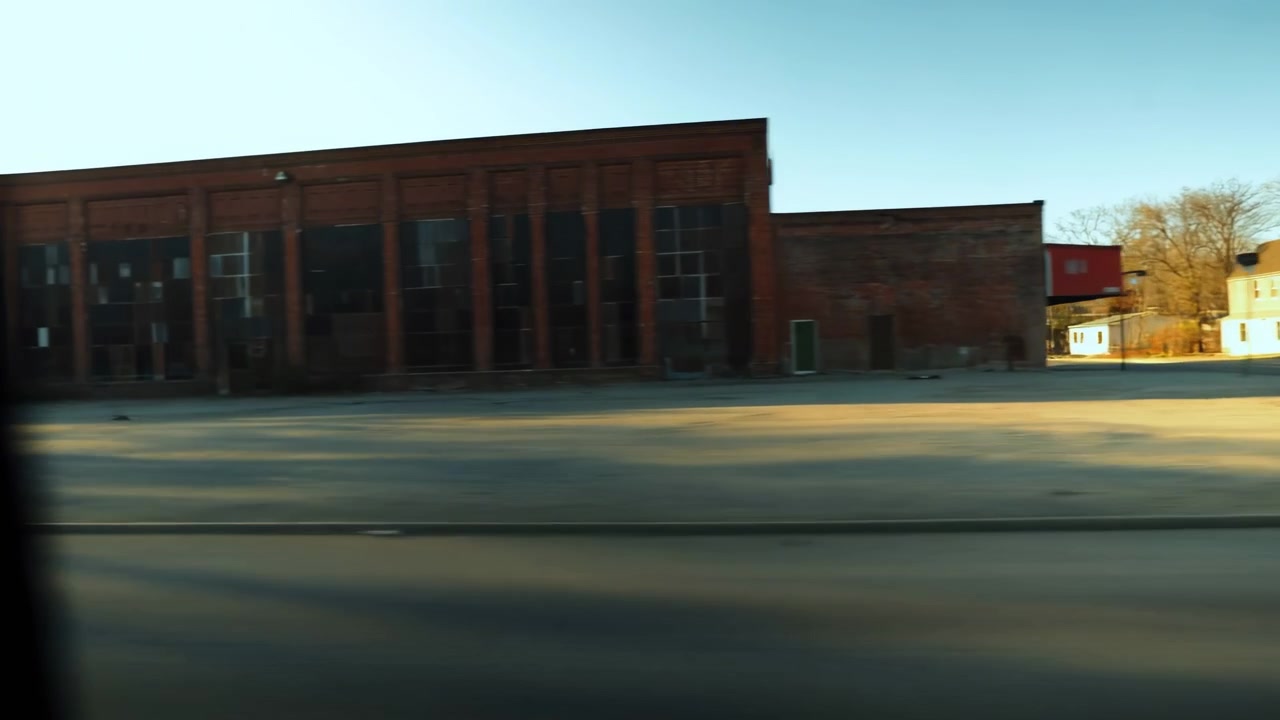 Calle soleada de pueblo pequeño pasando una fábrica de ladrillo rojo, vista borrosa desde la ventanilla del coche al borde de la carretera, ángulo bajo.