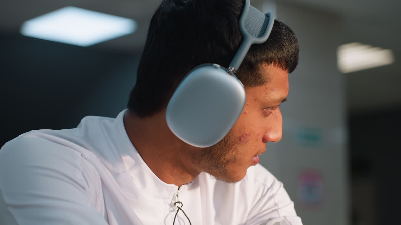 Close up of boy in white sports top wearing headphones, focused gaze toward blurred gym activity in background under bright ceiling lights and modern interior