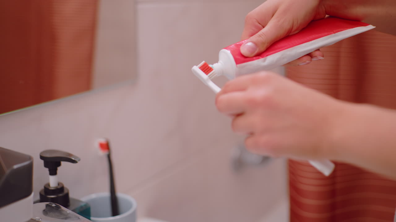 Close up person squeezing toothpaste onto toothbrush in bathroom sink area, showing morning hygiene routine, healthy lifestyle, dental care, personal grooming, and daily oral hygiene