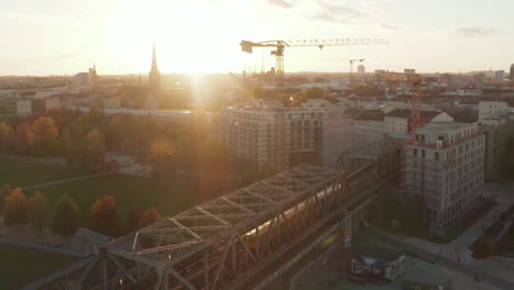 Gelber-Zug-Auf-Brücke-über-öffentlichem-Park-Mit-Menschen,-Die-Ihren-Sonnenuntergang-In-Berlin,-Deutschland,-Weitwinkelansicht-Aus-Der-Luft-Genießen