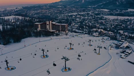 Stunning-Beautiful-Drone-Aerial-Hotel-and-Snow-Slopes-with-people-skiing-during-golden-hour