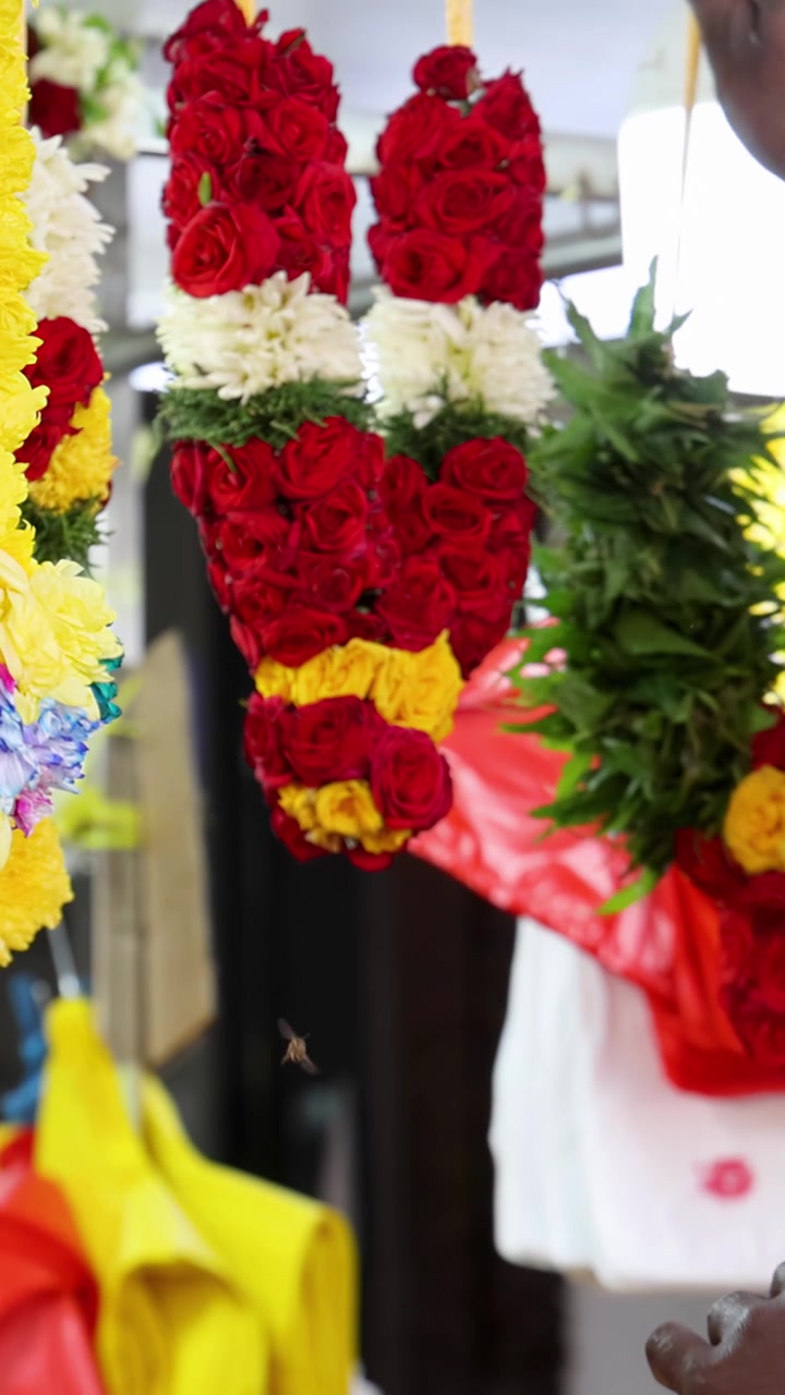 La mano di un venditore seleziona vivaci ghirlande di fiori da una colorata bancarella del mercato nella Little India di Singapore, sotto una luminosa illuminazione interna con bassa profondità di campo.