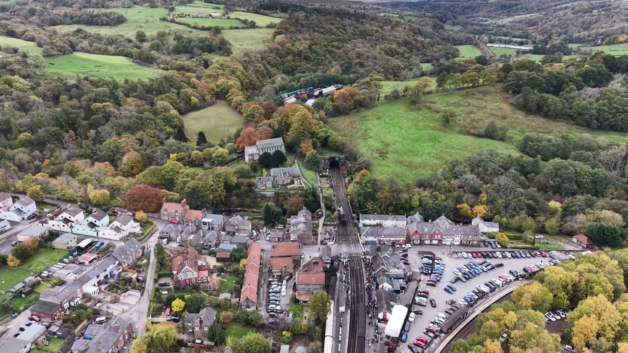 Aerial drone view of Pickering heritage railway station surrounded by autumn countryside in North Yorkshire, showing historic tracks, village buildings, cars, and fall foliage
