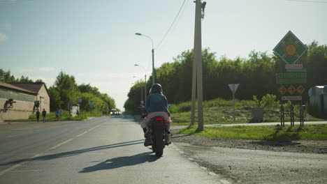 two ladies riding slowly on a power bike, approaching a road sign while cars come from the opposite direction, in the background, a few pedestrians walk along the sidewalk