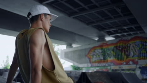 Sporty-teenager-resting-after-training-on-skateboard-at-skatepark-with-graffiti